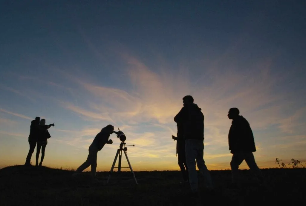 Silhouettes of people observing the twilight evening sky through a Newtonian telescope.